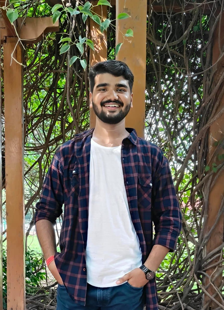 Adarsh Dwivedi smiling in a checked shirt in front of a vine-covered wall.